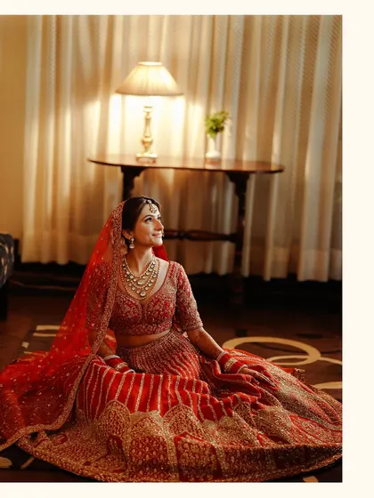 A beautiful indoor portrait of the bride sitting, her red lehenga spread around her. The warm lamplight creates a soft and intimate mood.