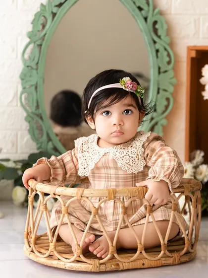 A calm and beautiful portrait of a baby girl in a plaid dress, seated in a rustic wicker basket before her cake smash.