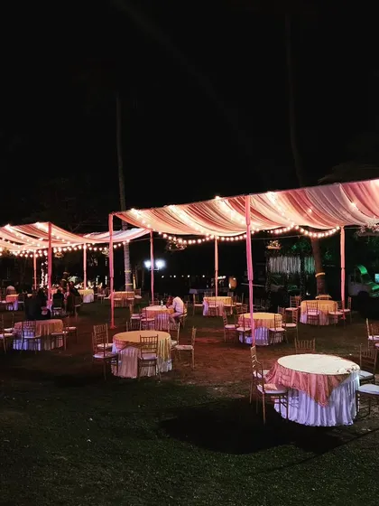 An outdoor cocktail setup featuring draped ceilings with vintage-style bulbs. This created a warm and inviting atmosphere for guests to mingle, with round tables set up on the lawn for a relaxed evening.