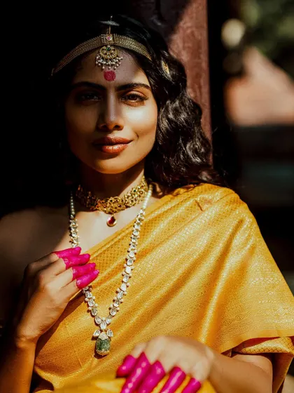 A close-up portrait from 'Thamara', focusing on the layered necklaces and the vibrant pink henna on the fingertips.