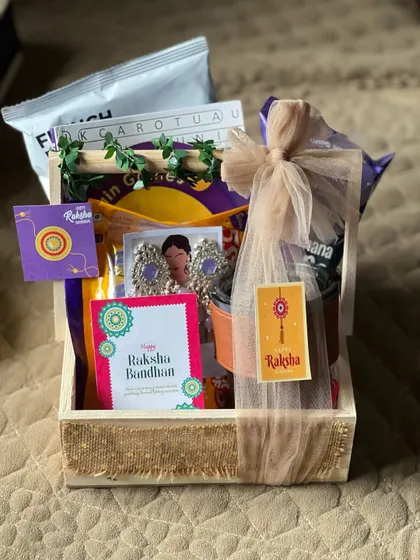A Rakhi hamper for a sister, featuring beautiful earrings, snacks, and a drink, all neatly arranged in a wooden crate.
