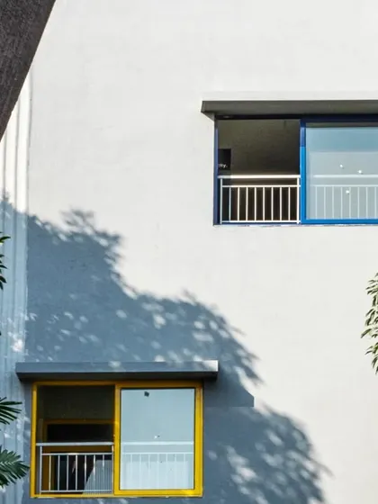 A close-up of the facade at L'Atelier, showing the contrast between the smooth plaster, colorful window frames, and the dappled light from nearby trees.