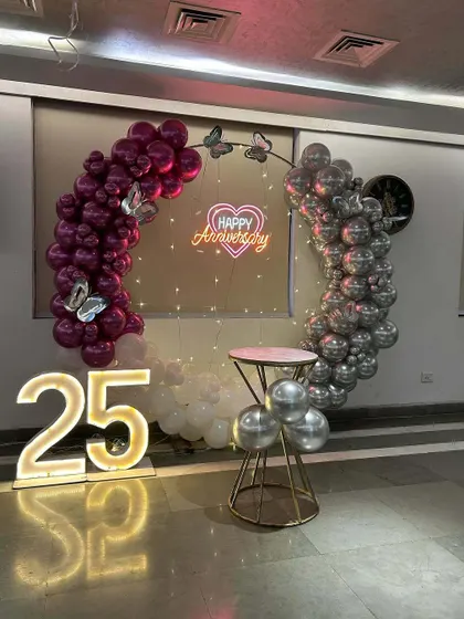 A romantic decoration for a 25th anniversary. This setup uses a circular frame with a split-color balloon design in maroon and silver, a 'Happy Anniversary' neon sign, and a '25' marquee number.