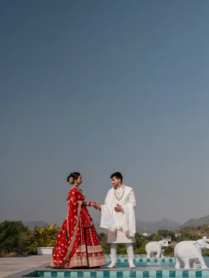 A majestic portrait of a couple by the poolside in Udaipur, with the Aravalli hills in the background. The composition emphasizes their regal attire and the luxurious setting of their destination wedding.
