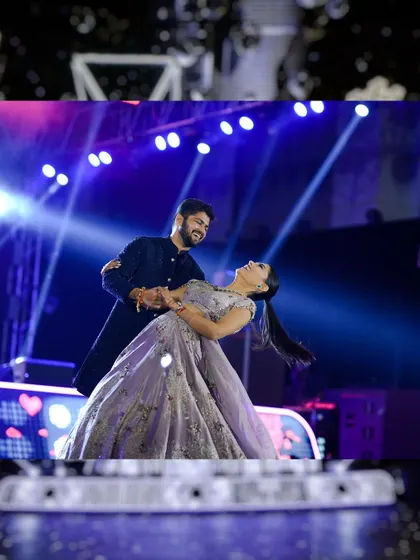 A beautiful moment from a couple's Sangeet dance. The groom dips the bride, who is wearing a flowing lavender gown, against a backdrop of bright stage lights, creating a picture-perfect romantic scene.