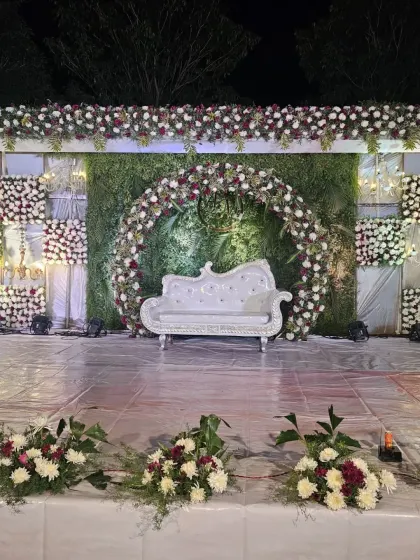 An outdoor reception setup featuring a stage with a lush green wall, a floral arch, and checkerboard floral panels for a textured look.