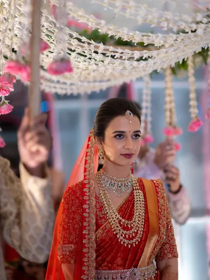 A beautiful South Indian bride in a red saree, making her entrance under a floral canopy.
