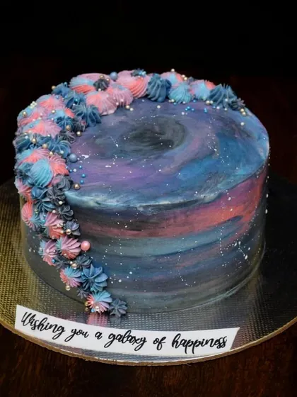 A galaxy-themed cake with a message "Wishing you a galaxy of happiness". The buttercream is painted in shades of blue, purple, and pink, with a swirl of piped stars on top.