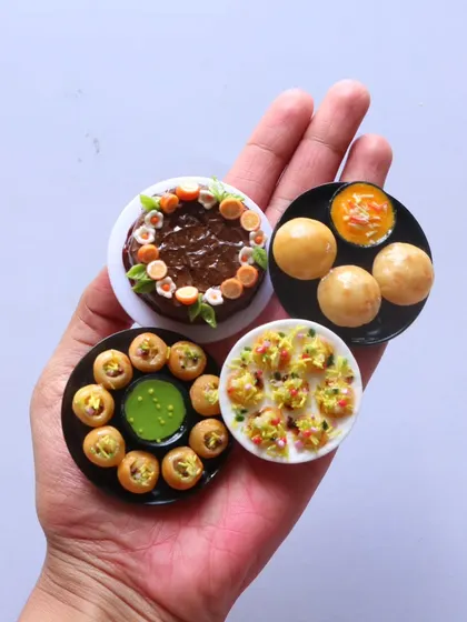 A hand holding a collection of chaat magnets, including pani puri, dahi puri, and a chocolate cake.