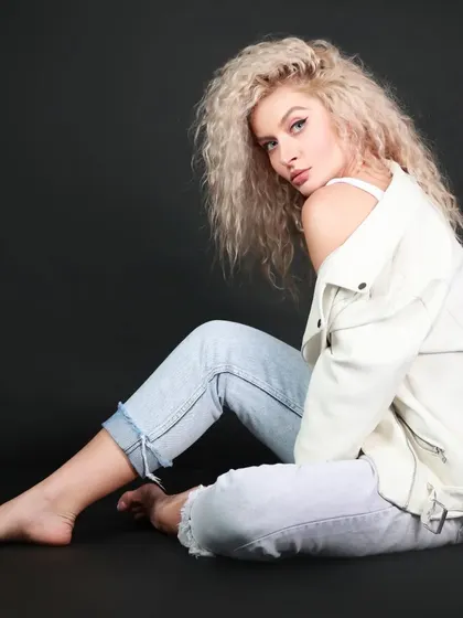 A full-body seated pose that showcases the model's long lines and relaxed confidence. The black background makes the light-colored outfit pop.