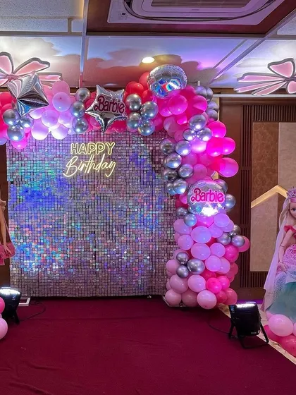 A Barbie-themed photo booth with a pink and silver balloon arch. The setup includes a shimmer wall, a neon 'Happy Birthday' sign, and life-sized Barbie doll cutouts.
