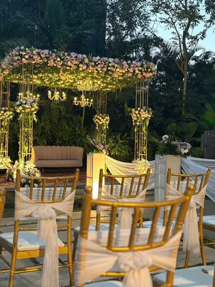 A romantic floral stage setup for an engagement, featuring a canopy of flowers, mini chandeliers, and elegant seating against a lush garden background.