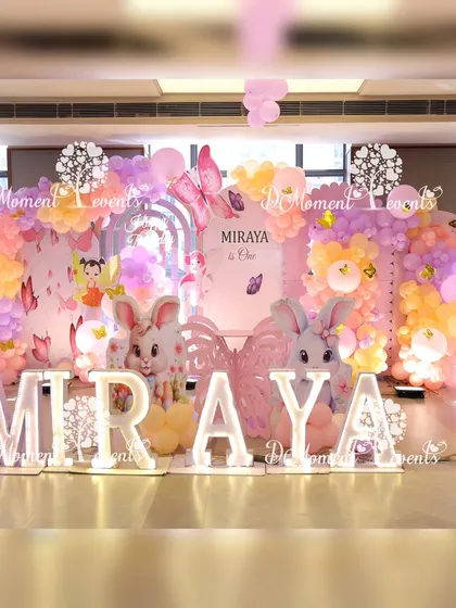 A magical stage setup combining butterfly, fairy, and bunny elements, with large bunny props and light-up letters for a whimsical first birthday.