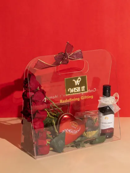 The "Love Affair" acrylic bag against a bold red background, highlighting its modern and chic design. It’s filled with Valentine’s essentials like roses, wine, and heart-shaped chocolates.