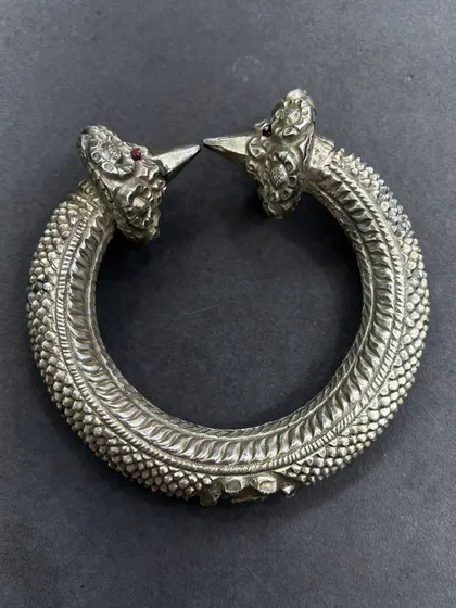 An exclusive silver leg kada, or anklet, with an antique finish. This piece features intricate scaling detail and two ornate heads with ruby-red stone eyes, a truly unique piece of tribal-inspired jewellery.