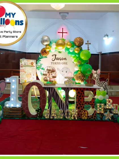 A wide shot of a jungle-themed first birthday inside a church hall. The setup features a large 'ONE' marquee table, animal cutouts, and a green and gold balloon arch.