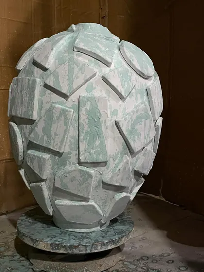 A large, textured globe vase after the first glaze application. The geometric shapes have been individually attached, and the light green celadon glaze is brushed on, ready for the transformative heat of the kiln.