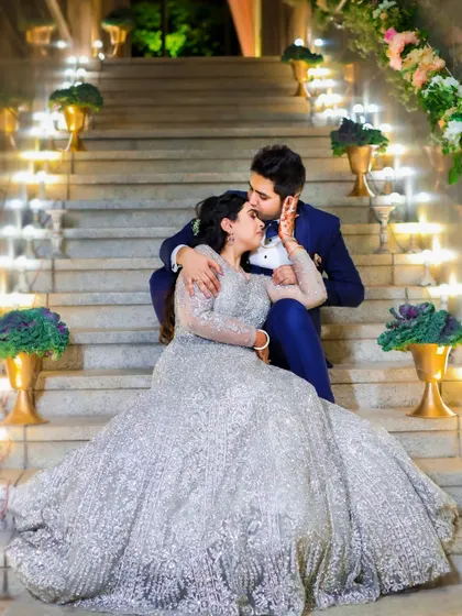 A fairytale-like portrait of the couple on a grand, lit-up staircase. The bride's voluminous silver gown and the groom's dapper blue suit create a scene of pure romance.