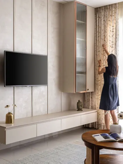 A minimalist TV unit in muted beige tones, featuring both closed and open storage. The design includes a floating shelf and a unique hanging cabinet with a fluted glass door and metallic accents.