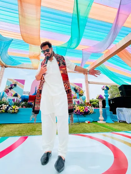 Mic in hand, addressing the crowd at an outdoor Mehendi event. The colorful drapes and bright sunshine create a perfect carnival atmosphere.