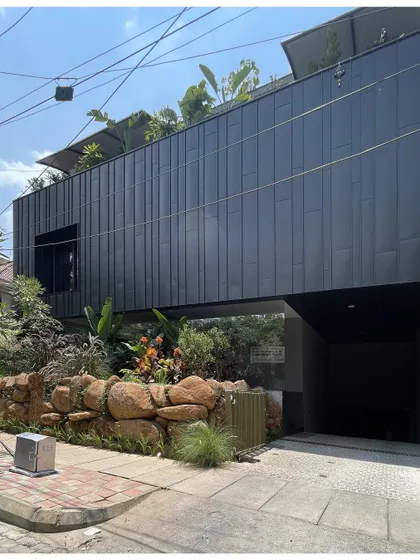 A view of a modern office block, where the upper volume appears to float above a landscaped ground floor. The design uses a cantilevered structure to create a dramatic architectural effect and a sheltered entryway.