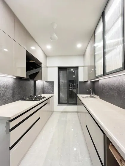 A modern parallel kitchen in a light grey finish. The design includes a long wall unit with a fluted glass shutter, a light quartz countertop, and a bold black textured backsplash for a chic, contemporary feel.