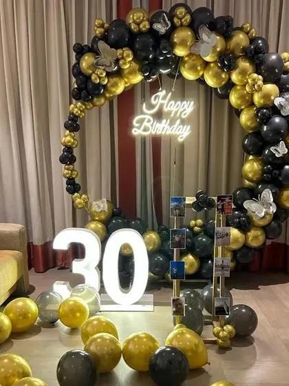 A 30th birthday party with a circular balloon arch in black and gold. The decoration includes a "Happy Birthday" neon sign, marquee numbers, and a photo display stand.