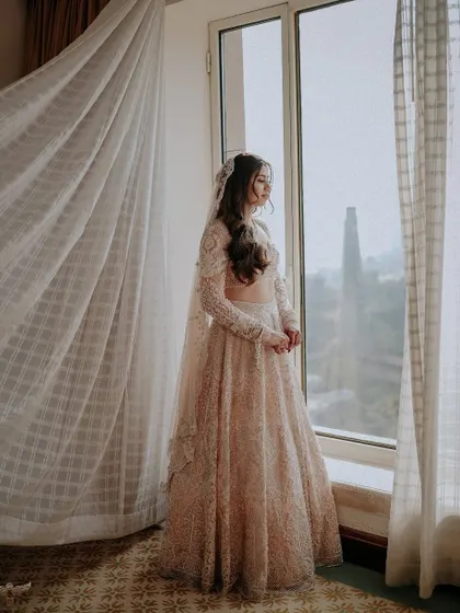 A pensive, window-lit portrait. The bride looks out, lost in thought, as the sheer curtain billows beside her, creating a soft, romantic, and cinematic feel.