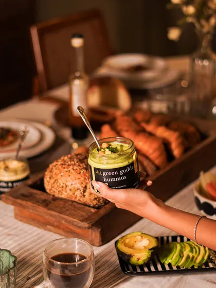 A jar of our vibrant Green Goddess Hummus, held over a beautiful breakfast spread. This creamy, herb-infused hummus is a healthy and delicious addition to any meal, perfect with fresh bread or vegetable sticks.