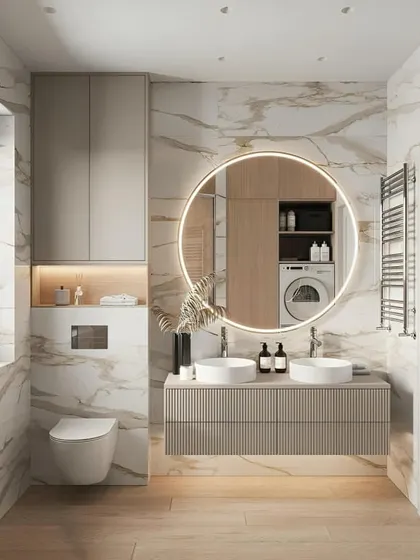 A bright and luxurious bathroom with marble walls and a floating beige vanity. The large round backlit mirror is a beautiful centerpiece, and the reflection shows a cleverly hidden laundry area.