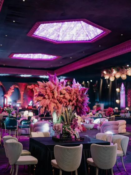 A dining table with a dramatic centerpiece of pink pampas grass, set under a ceiling with purple geometric light fixtures. The atmosphere is modern, moody, and luxurious.