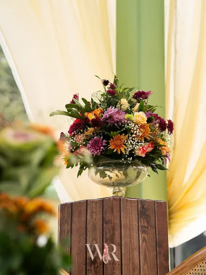 A large, vibrant floral arrangement sits atop a rustic wooden pedestal, set against soft, flowing drapes. This piece served as a beautiful focal point in the dining area.