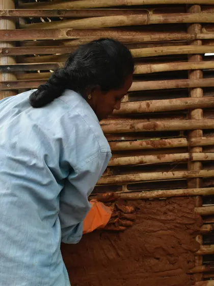 A woman mason applying the first layer of daub to a wattle wall. This is a key moment where the structure begins to transform into a solid, insulated partition.