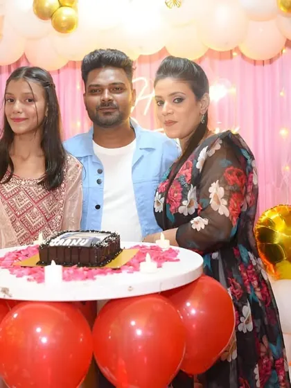 A family poses by the birthday cake table, surrounded by festive red and gold balloons.