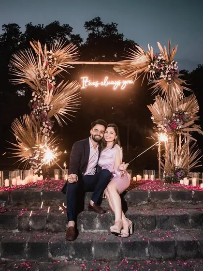 A wide shot of the entire proposal scene at night, with the floral arch, neon sign, and hundreds of candles creating a magical glow.