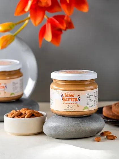 A balanced and serene composition featuring Being Better's almond butter. The product is placed on smooth grey stones, creating a natural, zen-like feel that aligns with a healthy lifestyle brand.