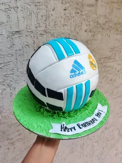 A fully spherical football cake, ready for a birthday celebration. The details are all hand-painted to create a realistic look, sitting on a field of green buttercream grass.