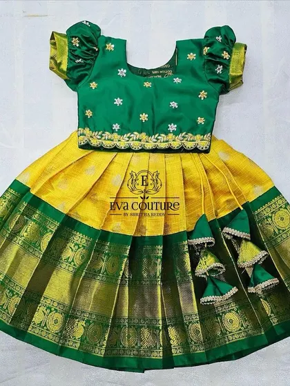 A cheerful yellow and green pattu pavadai for a toddler. The green blouse has simple white floral embroidery, and the skirt has a wide green and gold border.