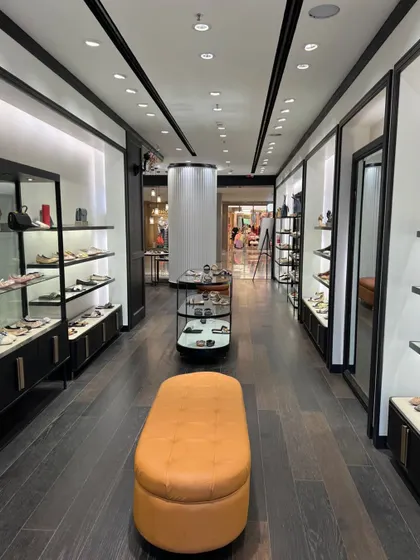 The main walkway of the Pazzion store, showing the clean layout, recessed wall displays with integrated lighting, and a central ottoman for customer seating.