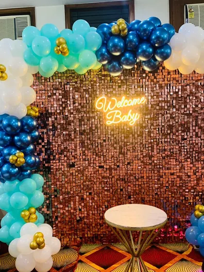 A "Welcome Baby" backdrop with a rose gold shimmer wall. The balloon arch in shades of blue, teal, and white with gold accents is perfect for welcoming a baby boy.