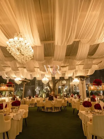 A wide view of the dining area, with its ceiling of draped white fabric and dozens of chandeliers. The tables are set with red floral centerpieces, creating a classic and romantic wedding reception.