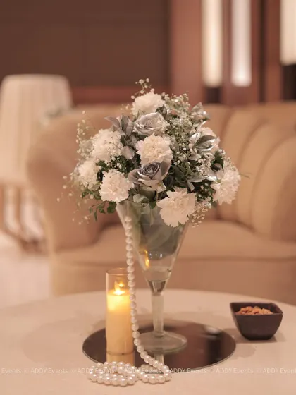 This centerpiece for a 75th birthday party features white and silver-sprayed flowers, a single candle, and a string of pearls. These elements combine to create a look of timeless elegance and celebration.