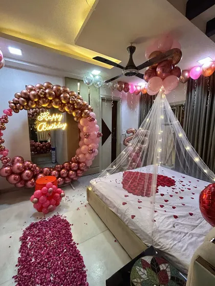 A wide view of a romantic birthday room setup. This decoration combines a large balloon ring, a bed canopy with lights, and extensive rose petal arrangements on the floor and bed for a grand romantic gesture.