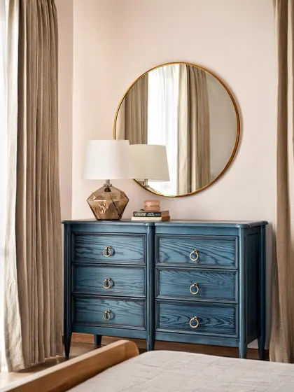 A stunning blue-stained dresser provides a beautiful pop of color against the soft blush walls of the primary bedroom. The round mirror and glass lamp add to the room's elegant, coastal feel.