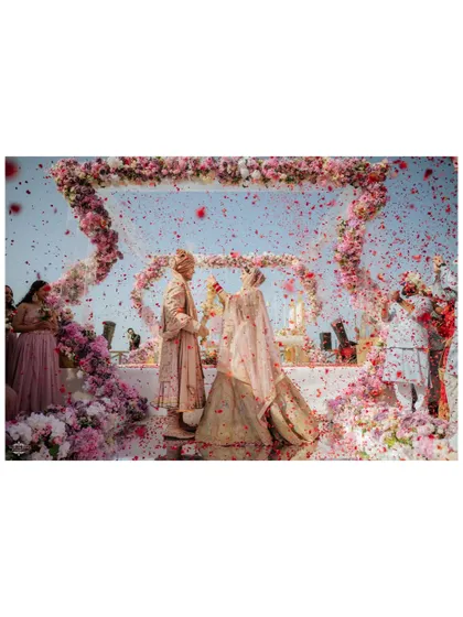 A wide shot of the Varmala moment, with a dramatic shower of pink and red petals against the blue sky.