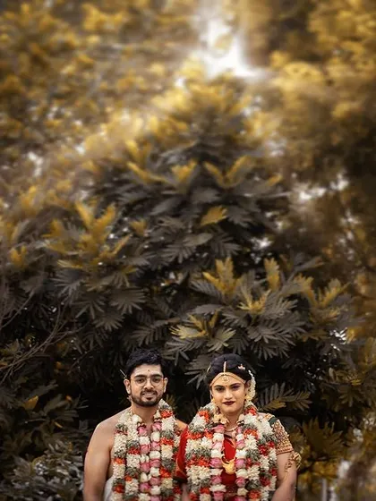 A formal portrait set against a backdrop of lush, golden-toned trees. This image showcases the couple adorned with garlands, a classic and beautiful memory from their wedding day.