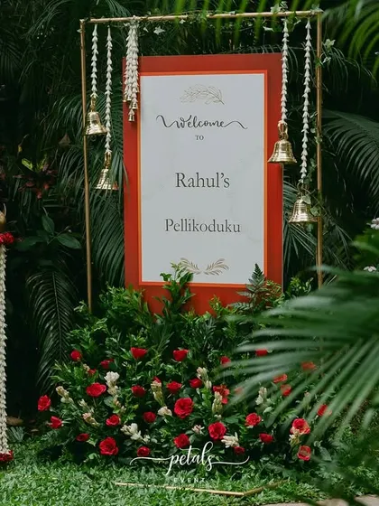 A welcome sign for a Telugu 'Pellikoduku' ceremony, framed in red and gold and adorned with hanging temple bells. The surrounding florals and traditional props add to the auspicious feel.