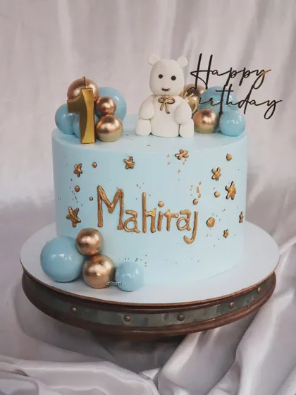 A sweet and simple first birthday cake for a little boy. This light blue cake features a cute teddy bear topper and is decorated with blue and gold spheres and little gold stars.
