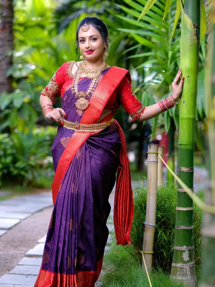 A full-length view of a bride in a purple and red Kanjivaram saree, set against a lush green background. The contrast of colors makes this a visually stunning choice for outdoor wedding photos.