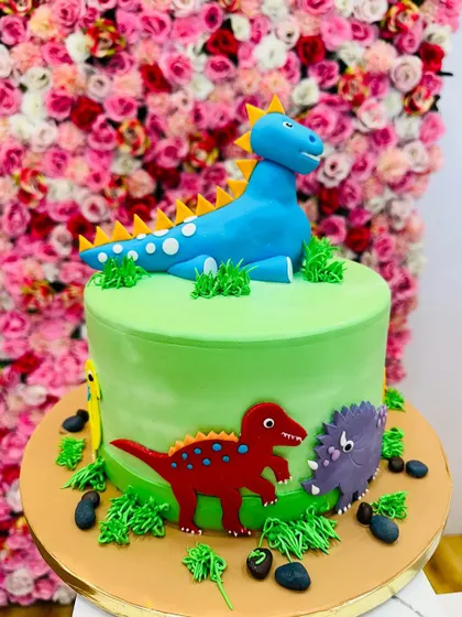 A fun dinosaur-themed cake with a large blue dinosaur on top and smaller dinos around the base. The green grass and edible rocks complete the prehistoric scene.